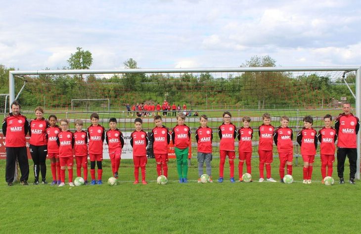 Eine Jugendfußballmannschaft in roten Trikots steht in Formation auf einem grünen Feld, neben ihnen ihre Trainer und im Hintergrund ein Tor.