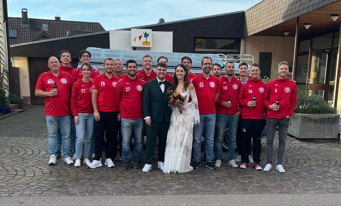 Eine Gruppe von Männern in roten Trikots posiert mit einer Braut in einem weißen Kleid, die in einer malerischen Umgebung im Freien feiert.