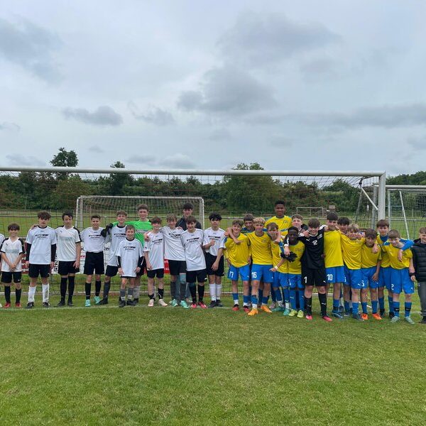 Zwei Jugendfußballmannschaften stehen gemeinsam lächelnd in weiß-gelben Trikots auf einer Wiese vor einem Torpfosten.