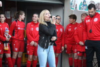 Eine Sängerin tritt bei einer Sportveranstaltung auf, während eine Frauenfußballmannschaft in roten Trikots ihr von hinten zusieht.