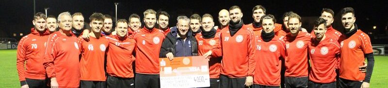 Eine große Gruppe von Menschen, meist Männer, in roter Sportkleidung, steht nachts auf einem Fußballfeld, lächelt und hält einen übergroßen Scheck in der Hand.