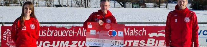 Drei Personen in roten Jacken stehen im Freien auf einem verschneiten Feld und halten einen großen Scheck in der Hand. Der Scheck ist auf 3000 Euro ausgestellt und stammt von einer Brauerei. Die Szene ist hell und fröhlich.