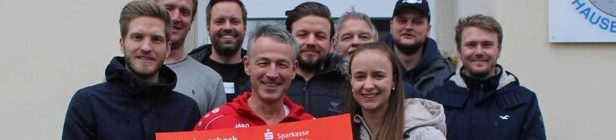 Eine Gruppe von Menschen, meist Männer und zwei Frauen, steht im Freien und hält einen großen roten Scheck in der Hand. Sie lächeln und scheinen glücklich zu sein. Der Schauplatz befindet sich vor einem hellen Gebäude. Die gesamte Farbpalette ist gedämpft, wobei der rote Scheck die auffälligste Farbe ist.