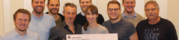 Eine Gruppe von Männern und eine Frau lächeln für ein Foto, während sie einen großen überdimensionalen Scheck halten. Sie stehen in einem Innenraum vor einer hellbeigen Wand. Die allgemeine Stimmung ist fröhlich und feierlich.
