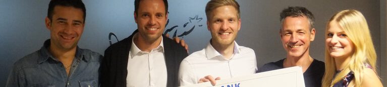 Ein Team von Menschen, die freudig einen Scheck in der Hand halten und vor einer lebendigen Wand voller Kunstwerke stehen.