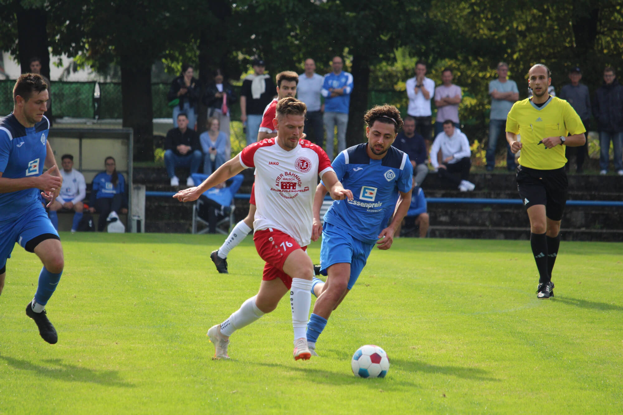 Ein dynamischer Moment während eines Fußballspiels, bei dem Spieler in roten und blauen Trikots auf einem grünen Feld in Aktion sind, mit Zuschauern im Hintergrund.