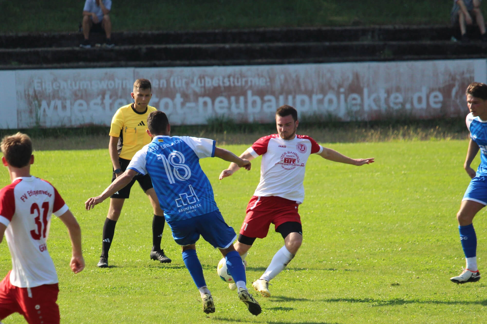 Ein Fußballspiel im Gange, zwei Spieler in Aktion mit einem Schiedsrichter, der beobachtet. Das Spielfeld ist leuchtend grün, im Hintergrund sind Zuschauer zu sehen.