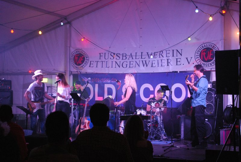 Eine quirlige Band spielt auf der Bühne unter bunten Lichtern, im Hintergrund hängt ein Banner mit der Aufschrift „Fußballverein Ettlingenweier E.V.“.