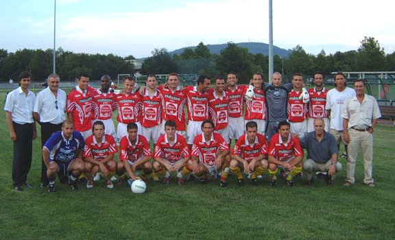 Eine Fußballmannschaft posiert in roten Trikots auf einer Wiese, umgeben von Trainern und Mitarbeitern, mit Bergen im Hintergrund.