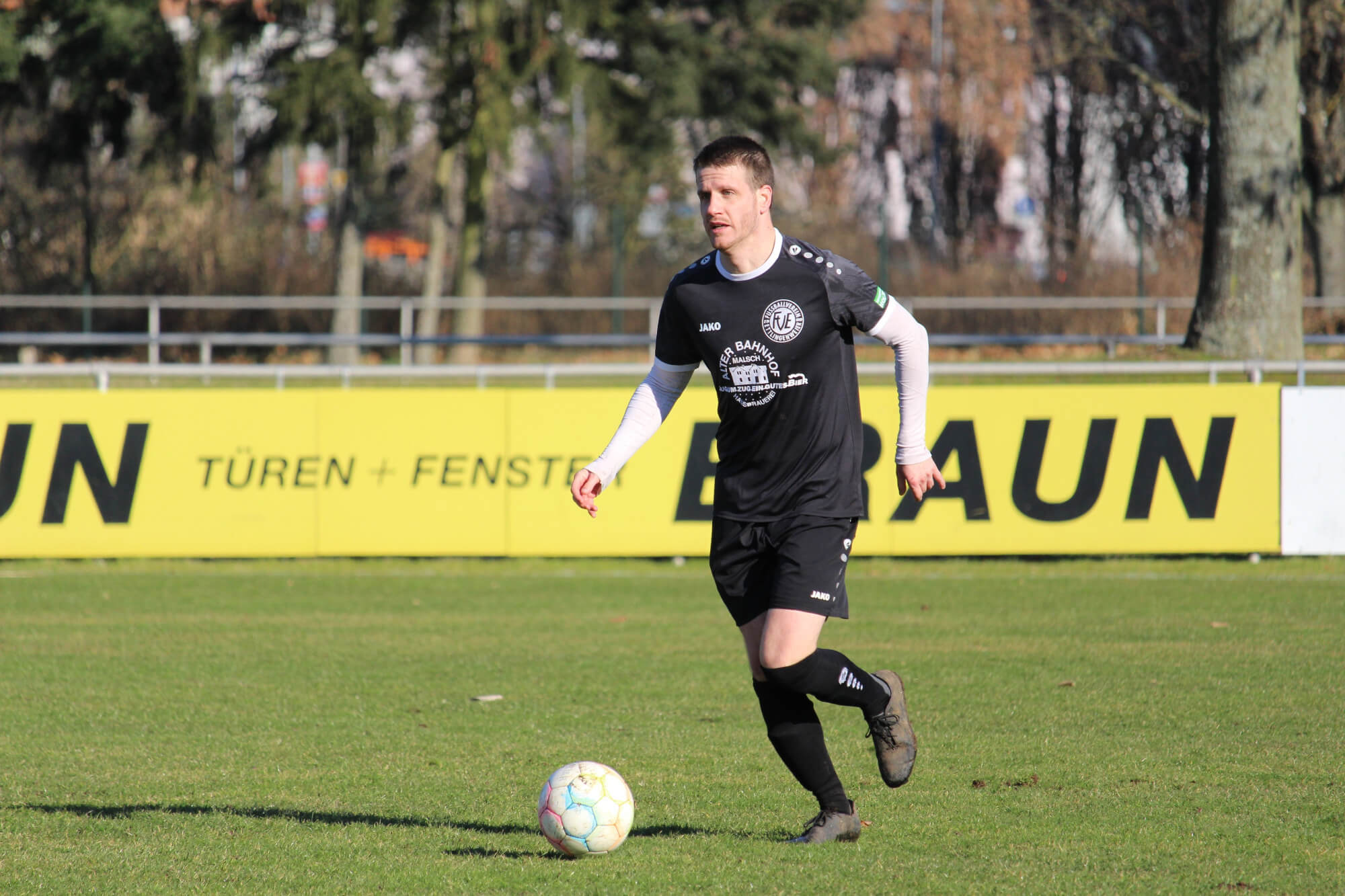 Ein Fußballspieler in schwarzer Ausrüstung, bestehend aus Trikot und Shorts, dribbelt einen Ball auf einem Rasenfeld. Unter dem Trikot trägt er weiße Ärmel. Im Hintergrund ist ein gelbes Werbeschild zu sehen. Bäume und ein Zaun sind ebenfalls in der Szene zu sehen.
