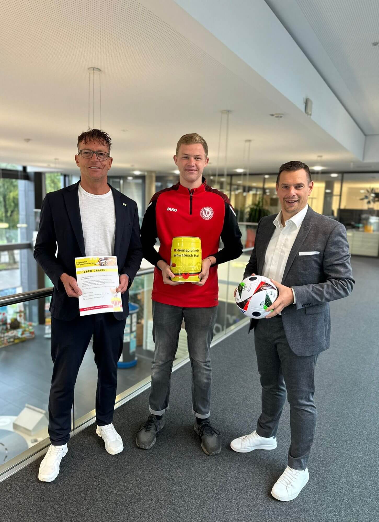 Drei Männer stehen in einer modernen Büroumgebung und halten einen gelben Container, einen Flyer und zwei Fußbälle in der Hand, um für eine lokale Veranstaltung zu werben.