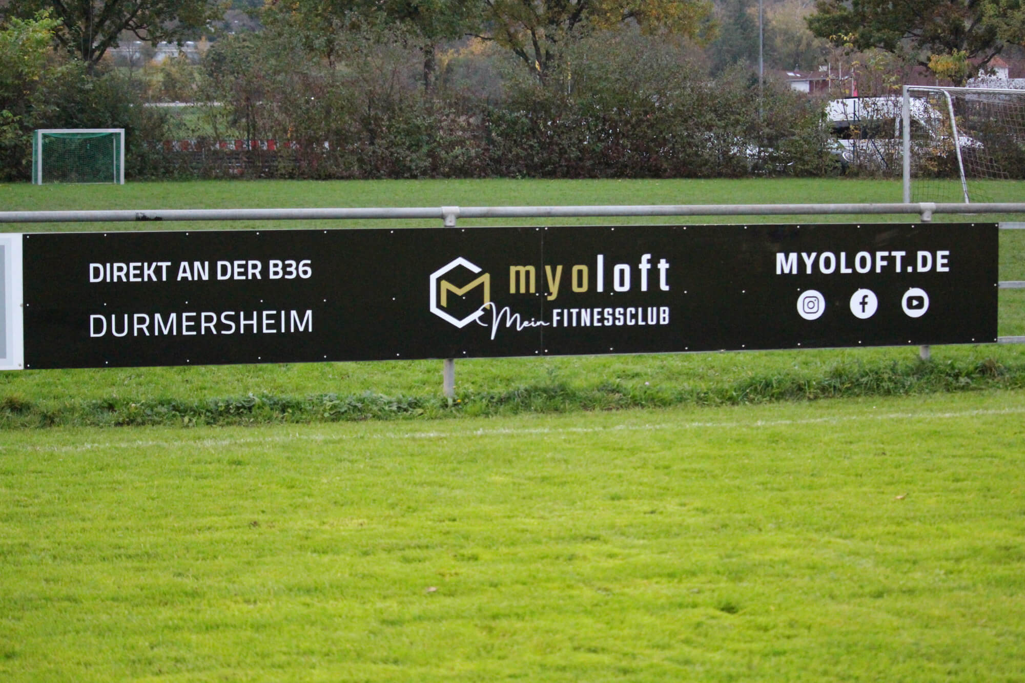 Ein schwarzes Werbebanner für den Fitnessclub „myoloft“ in Durmersheim, das auf einem Sportplatz angebracht ist und Kontaktsymbole für soziale Medien enthält.