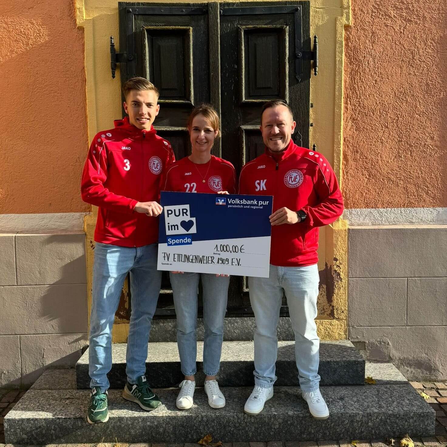 Drei Personen in roten Sportjacken halten einen großen Scheck über 1.000 Euro zur Unterstützung des FV Eitlingenweiler 1909 e.V. auf einer Steinstufe.