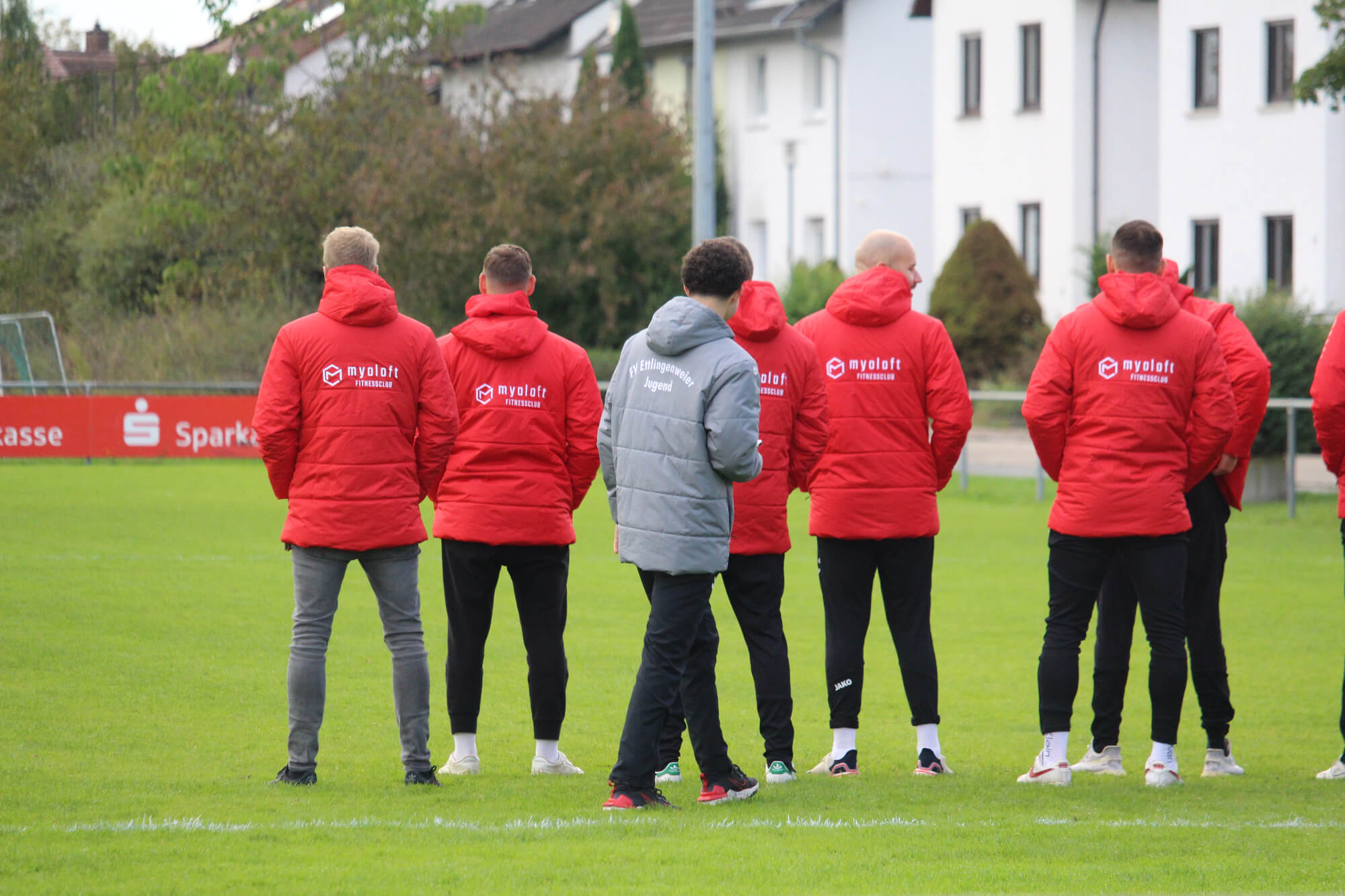 Eine Gruppe von Trainern in roten Jacken beobachtet eine Trainingseinheit auf einem grünen Feld, mit weißen Gebäuden und Bäumen im Hintergrund.