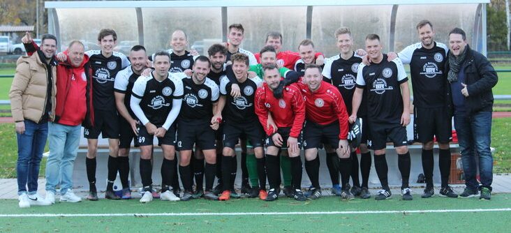 Eine Gruppe von 14 Fußballspielern in schwarzen Trikots und Shorts posiert gemeinsam auf dem Spielfeld und feiert vor der Spielerbank einen Mannschaftsmoment.