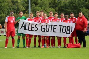 Die Startelf des FVE im Spiel gegen die SG Stupferich hält vor dem Anpfiff ein Banner mit der Aufschrift "Alles Gute Tobi"