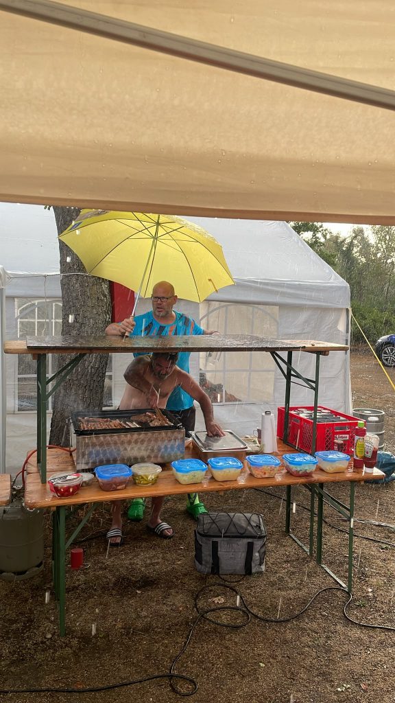 1 Mann grillt und ein anderer Mann hält einen Schirm darüber, da es stark regnet